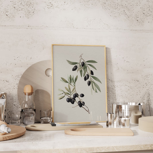Framed olive botanical art print in a natural oak frame, styled on a modern kitchen counter with wooden boards, glassware, and stainless steel accents.