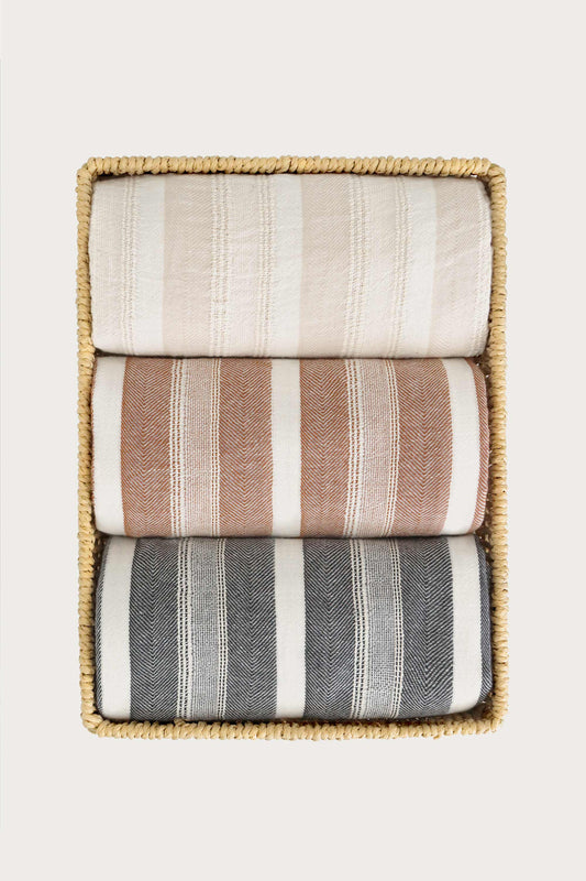 Avery Home Formentera striped peshtemal Turkish towels in sand, terracotta, and black herringbone, folded and stacked in a woven basket, Turkish cotton hammam towel gift set