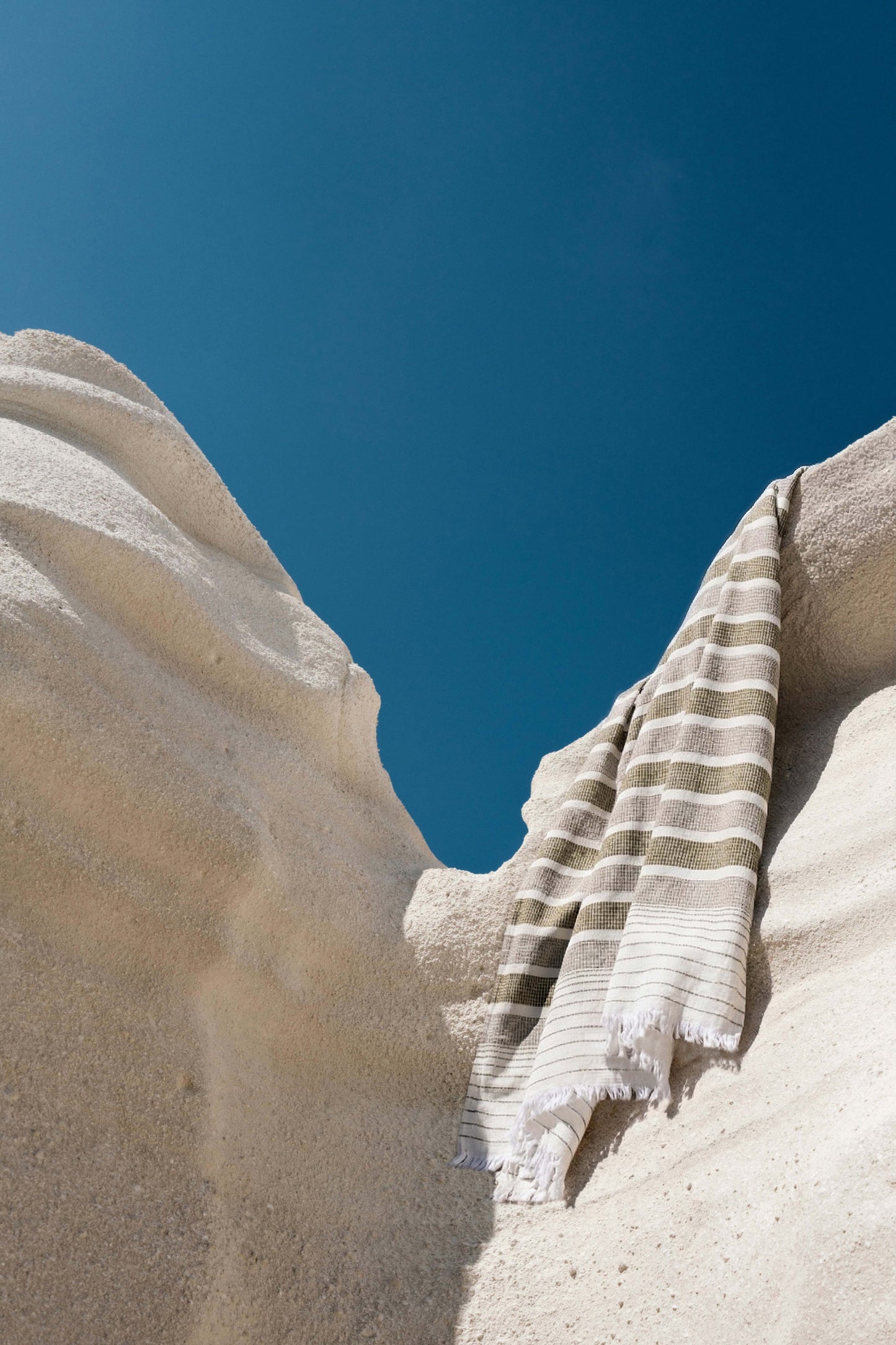 Avery Home olive stripe peshtemal draped over sun-bleached Mediterranean rocks against deep blue sky, lightweight Turkish hammam towel for beach and travel