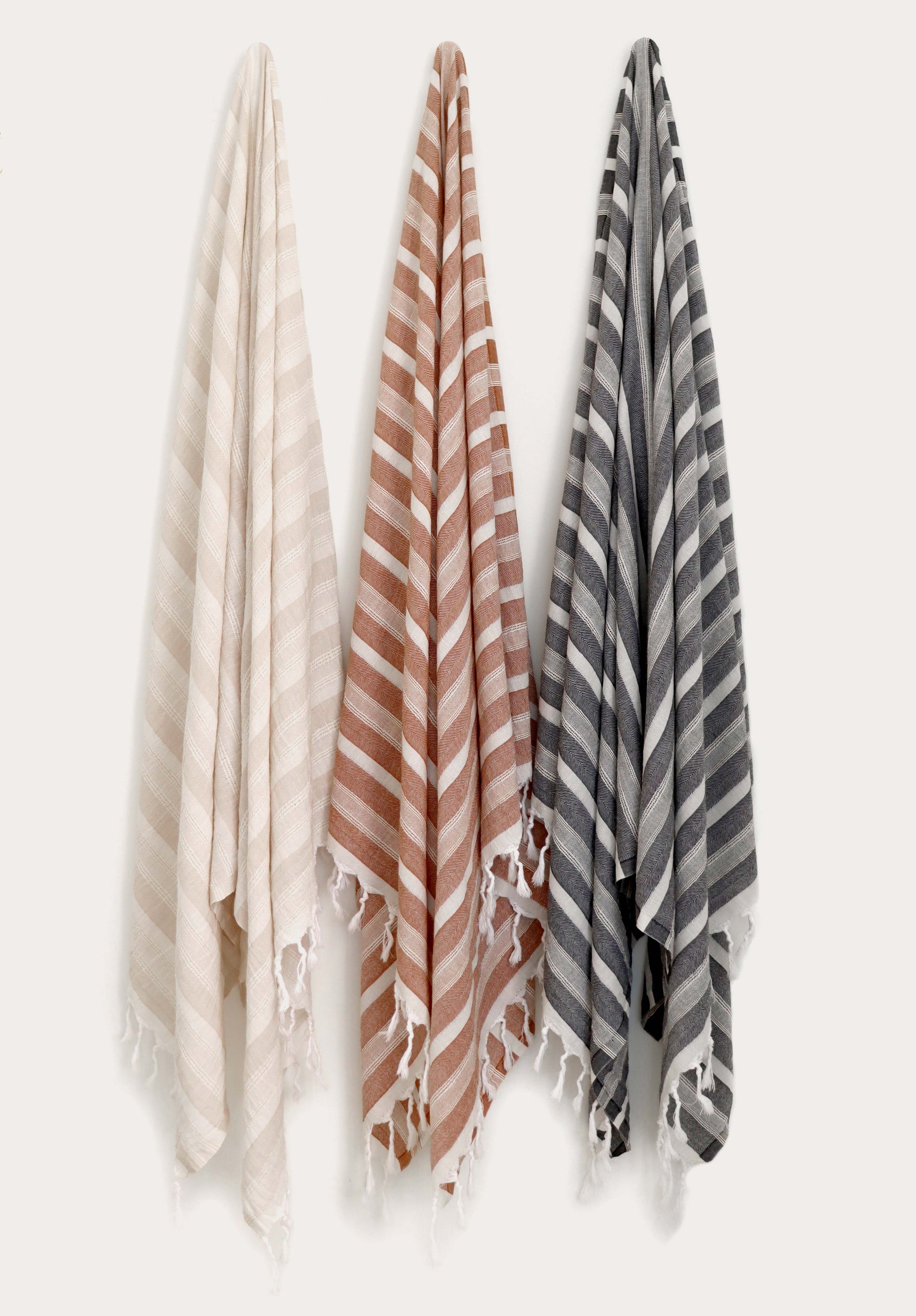 Collection of three flat weave Turkish cotton towels in beige, terracotta, and anthracite hanging on wall hooks. Soft, lightweight, quick-drying Turkish towels with hand-tied tassels for beach, bath, spa, travel, and everyday use by Avery Home