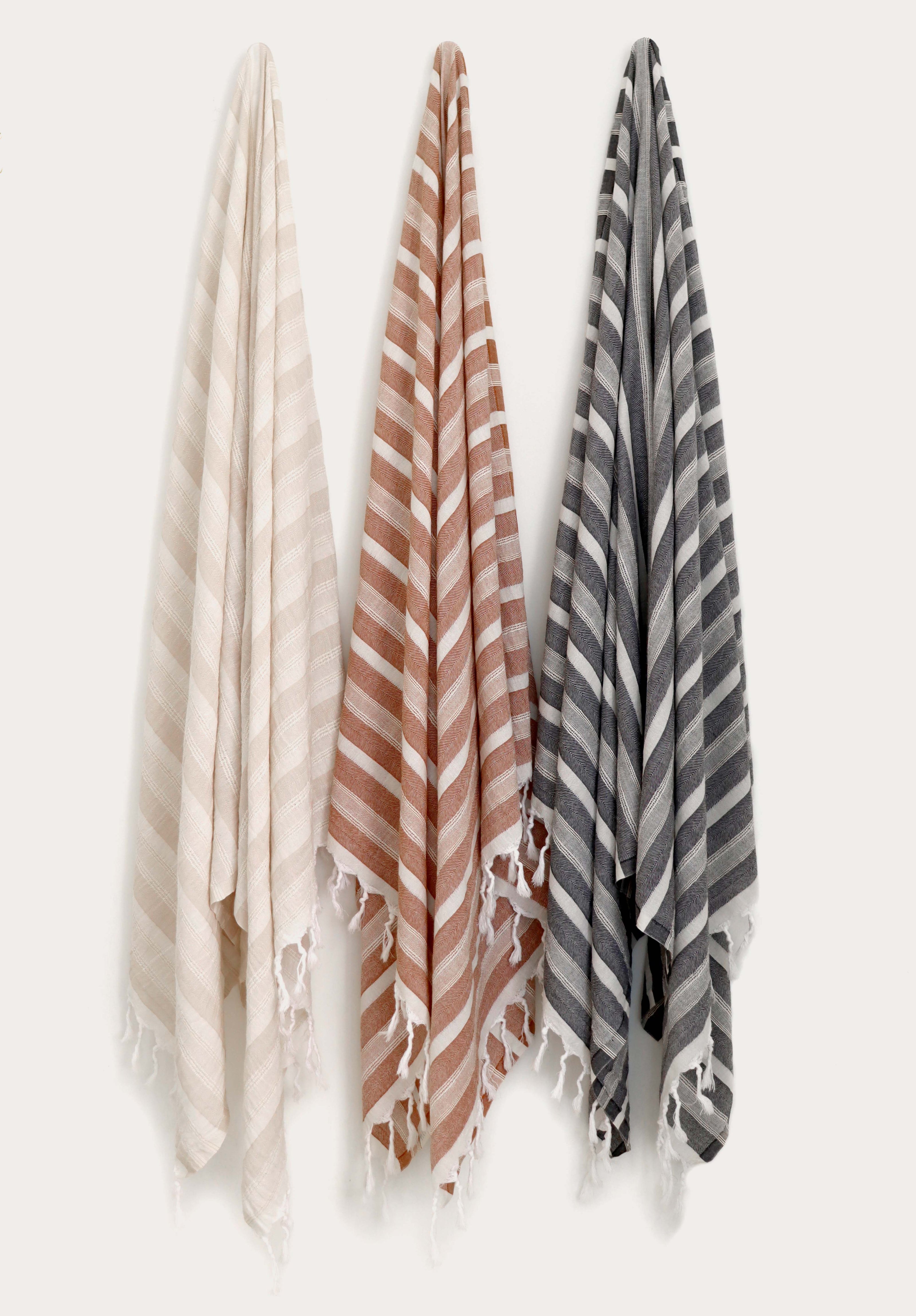Collection of three flat weave Turkish cotton towels in beige, terracotta, and anthracite hanging on wall hooks. Soft, lightweight, quick-drying Turkish towels with hand-tied tassels for beach, bath, spa, travel, and everyday use by Avery Home