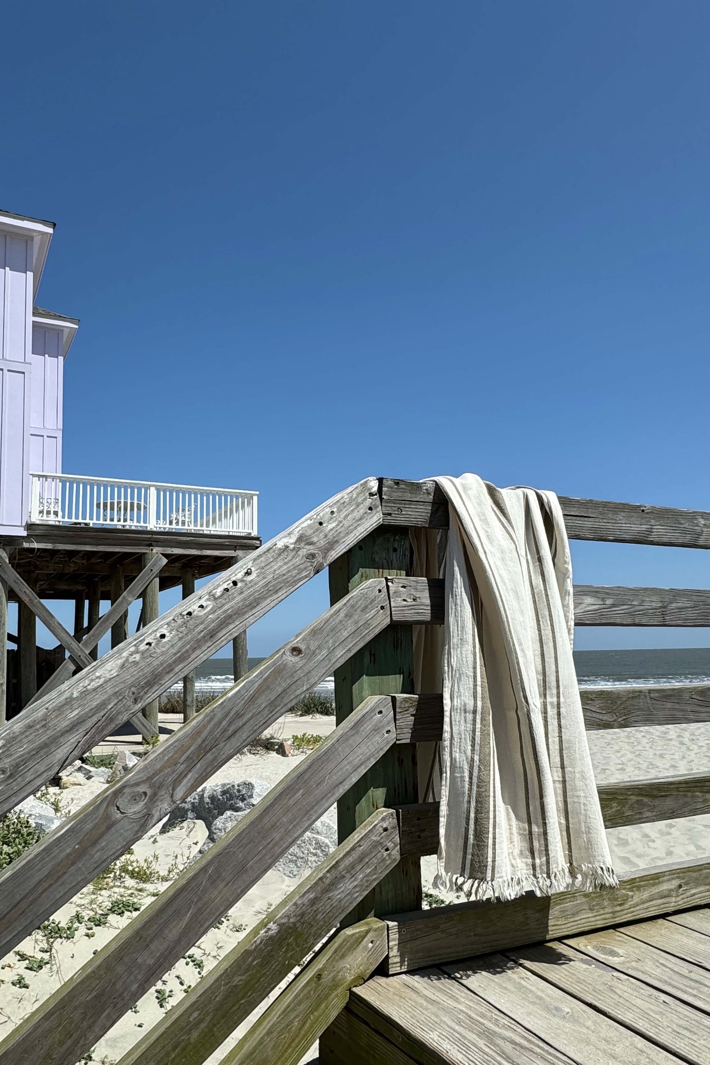 Green striped Turkish cotton towel draped over a weathered wooden beach boardwalk railing overlooking sandy dunes and the ocean under a clear blue sky. Lightweight, quick-drying Turkish towel for beach, travel, pool, and coastal living by Avery Home.