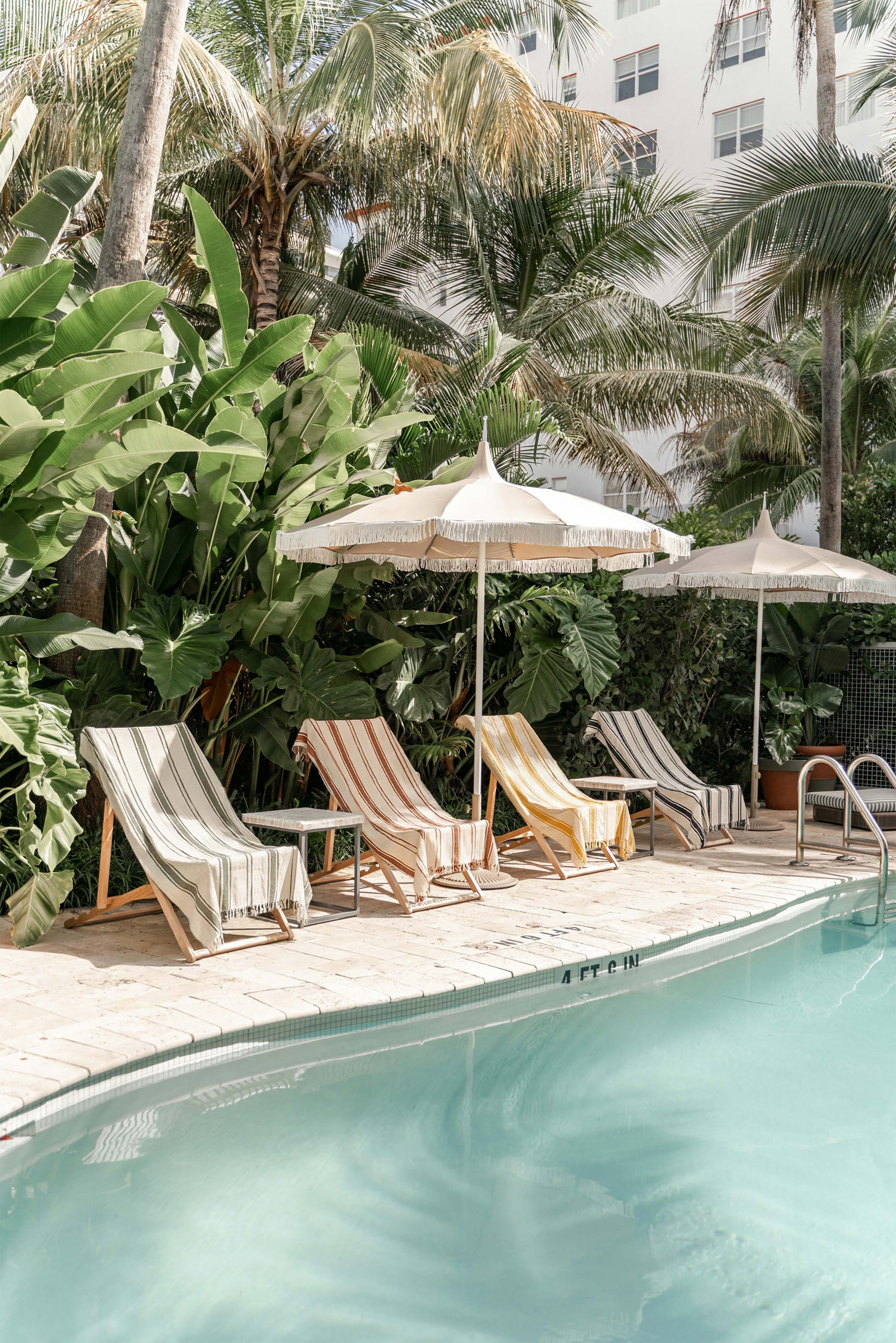 Striped cotton Turkish towels draped over wooden poolside lounge chairs at a luxury resort, surrounded by tropical palm trees — shop beach towels, pool towels, and Turkish towels