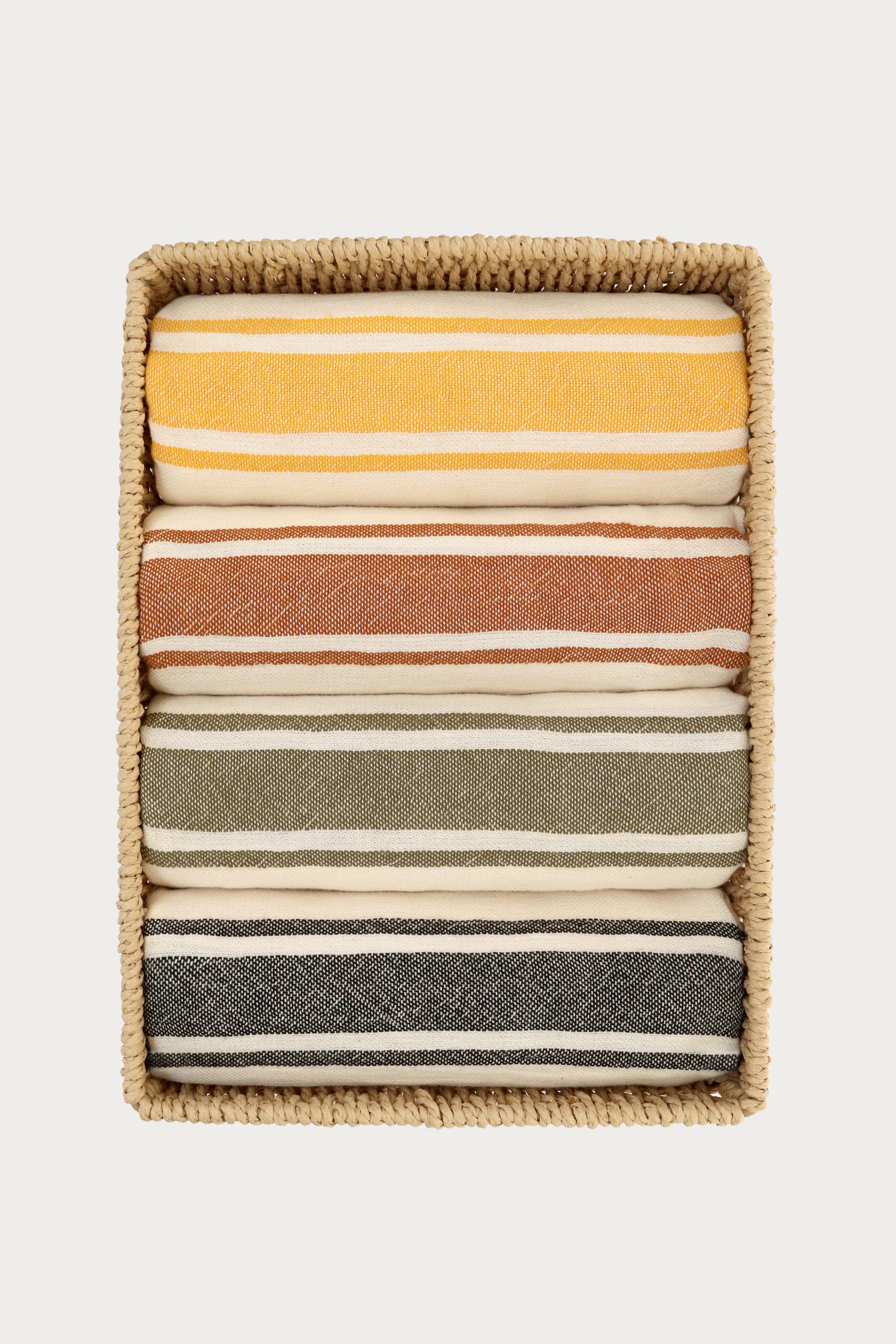 Set of four striped Turkish towels in yellow, terracotta, green, and black neatly folded inside a woven storage basket. Lightweight, absorbent Turkish cotton towels for beach, bath, travel, and home décor by Avery Home.