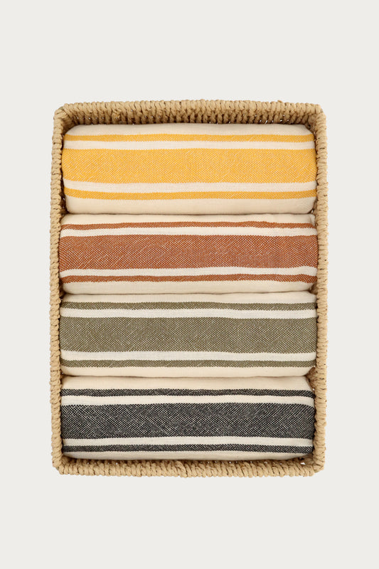 Set of four striped Turkish towels in yellow, terracotta, green, and black neatly folded inside a woven storage basket. Lightweight, absorbent Turkish cotton towels for beach, bath, travel, and home décor by Avery Home.